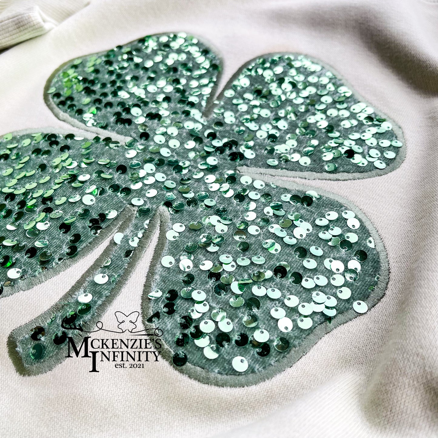 Sequined four-leaf clover design on a light green background with 'Mckenzie Infinity' branding.