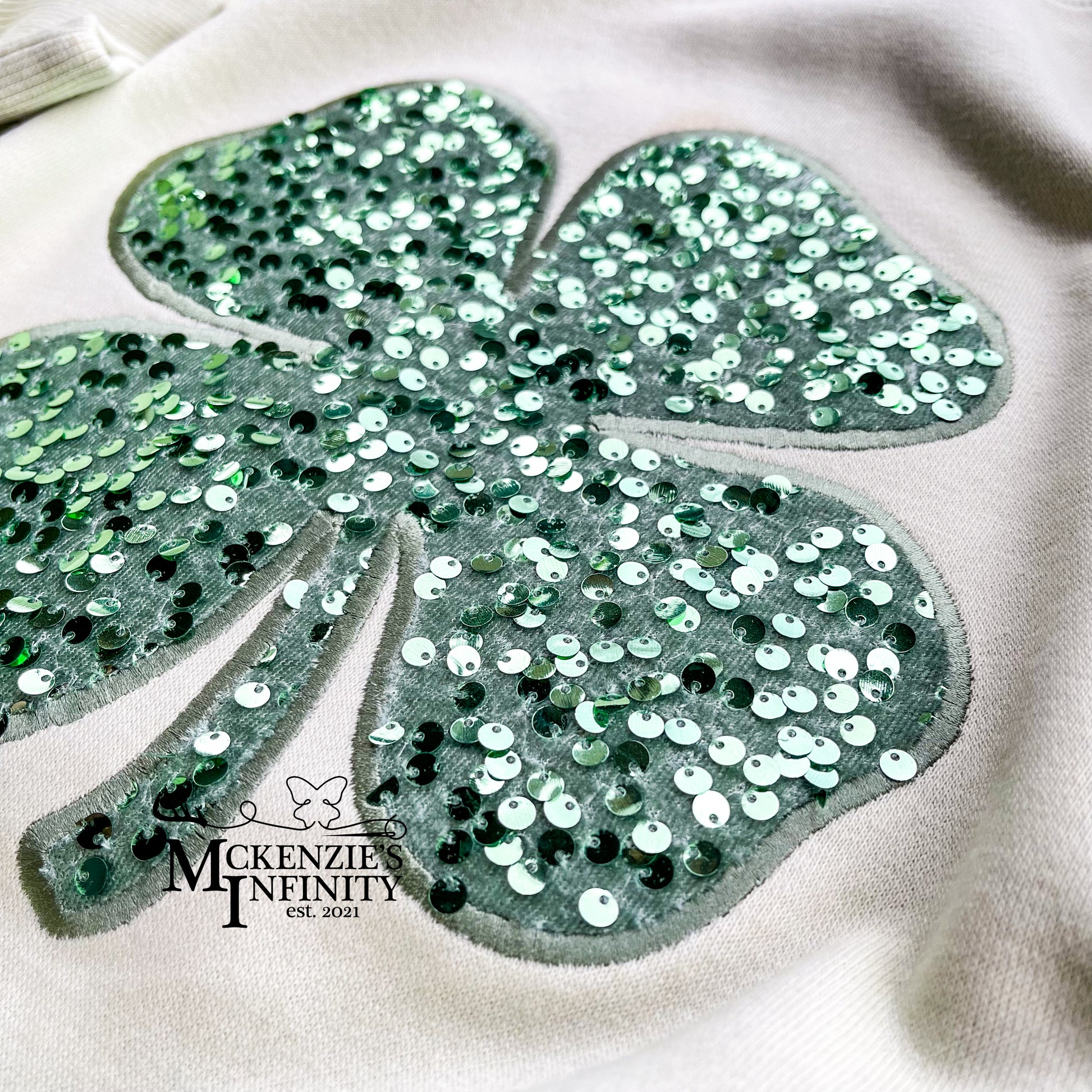 Sequined four-leaf clover design on a light green background with 'Mckenzie Infinity' branding.
