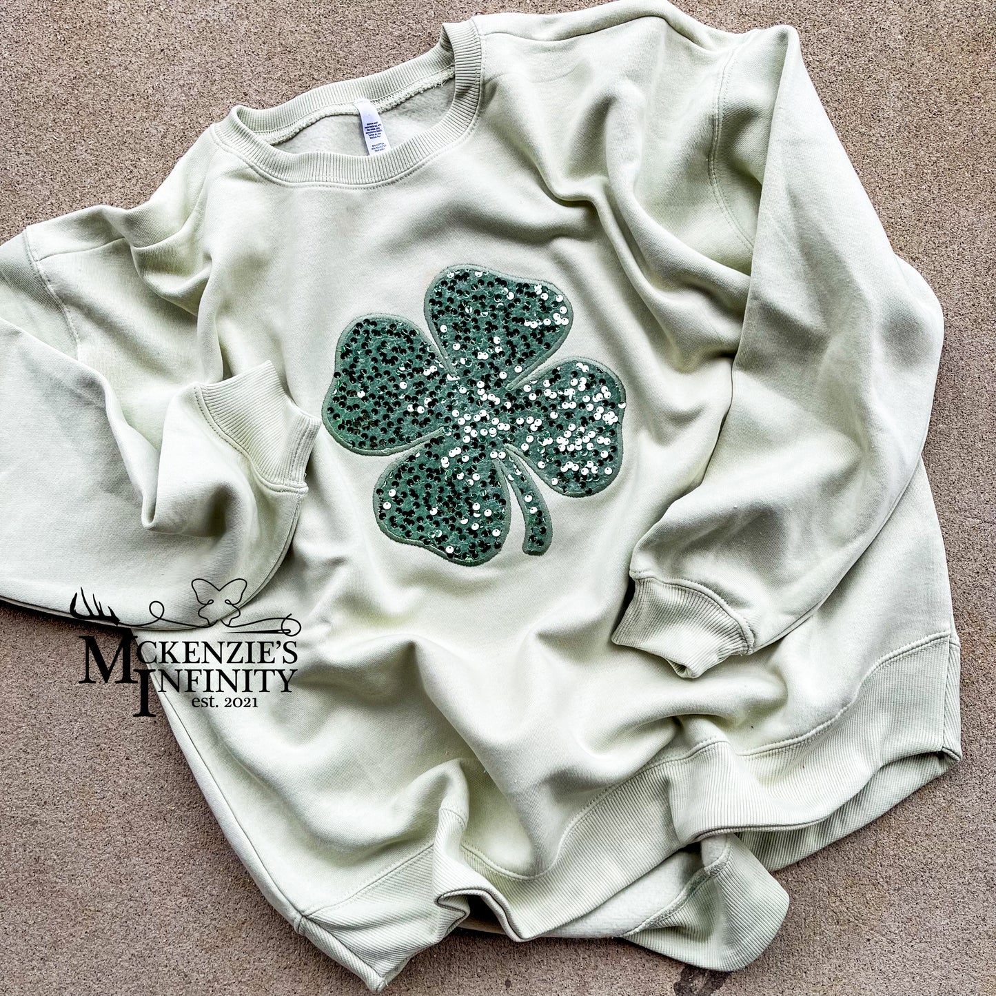Light green sweatshirt with a green sequin shamrock design on a beige surface, branded 'Mckenzie's Infinity'.
